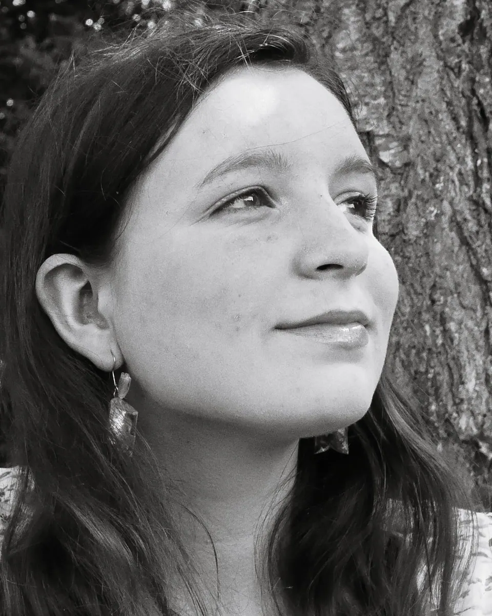 Black-and-white portrait of Alexandra: she smiles and looks up and away from the camera, with long dark hair and dangling earrings, in front of a tree trunk.